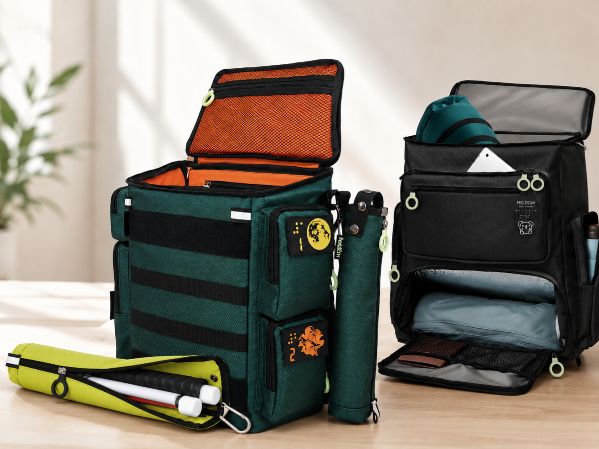 Feeldom disability bags and cane holsters displayed on a table, including an open backpack, black travel bag, and kiwi green cane pouch holding a folded white cane.